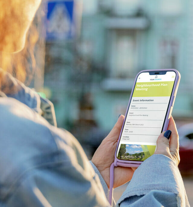 Young red haired woman holding an iphone with the screen of the community engagement app showing a Neighbourhood Plan meeting screen