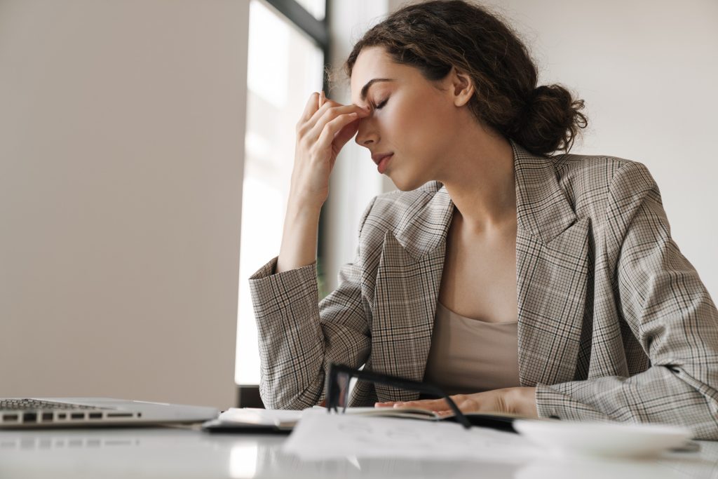 Stressed professional woman, arms resting on the desk, one hand holding the bridge of her nose with her eyes closed